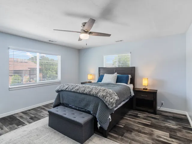 Primary bedroom featuring wood-finish flooring, two window openings with blinds, a ceiling fan with integrated lighting, and a dark wood-finish headboard