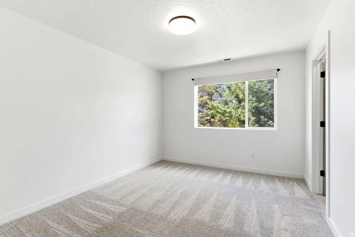 Bright interior room featuring a window with roller shade, white trim, light-toned patterned carpet, flush-mount ceiling lighting, and a white interior door with black hardware