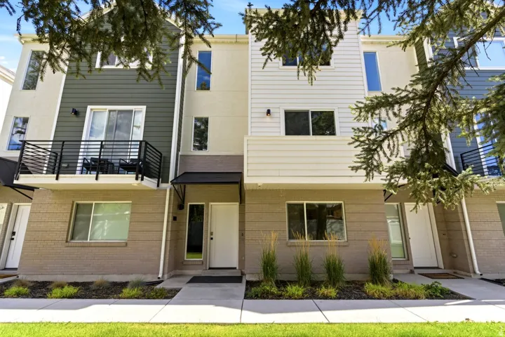 Modern townhome exterior featuring an upper-level balcony with black metal railings, a contemporary awning over the entrance, and a light-colored brick facade on the lower level
