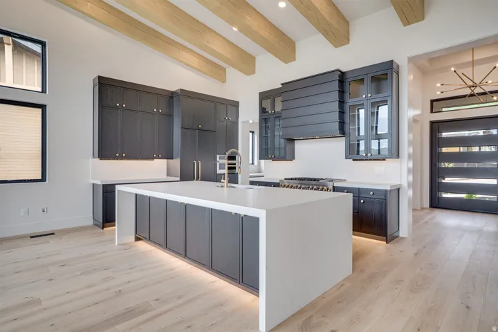 Open-concept kitchen featuring a large waterfall island with integrated lighting, dark wood-finish cabinetry, light wood-finish flooring, and exposed ceiling beams