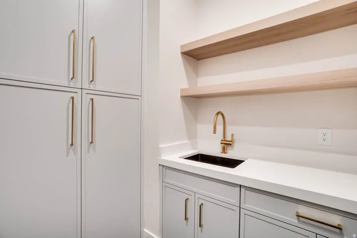 Custom cabinetry with gold-finish hardware, a built-in sink with a gold-finish faucet, white countertops, and wood-finish floating shelves