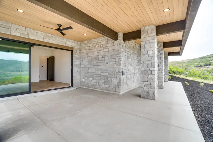 Spacious concrete patio featuring a natural stone facade, wood-finish ceiling with exposed beams, recessed lighting, and a ceiling fan