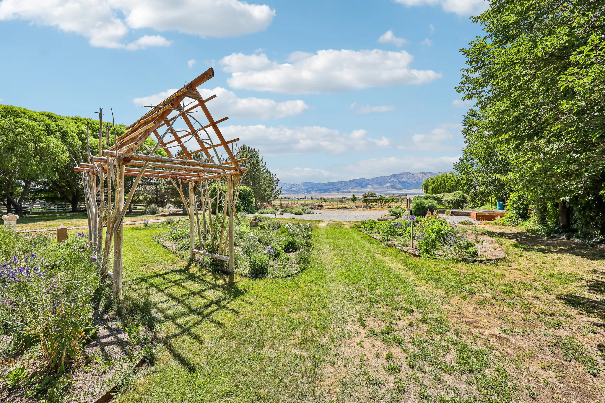 Expansive outdoor space featuring a rustic wood arbor, verdant lawn, mature trees, and garden beds, all set against a distant mountain range
