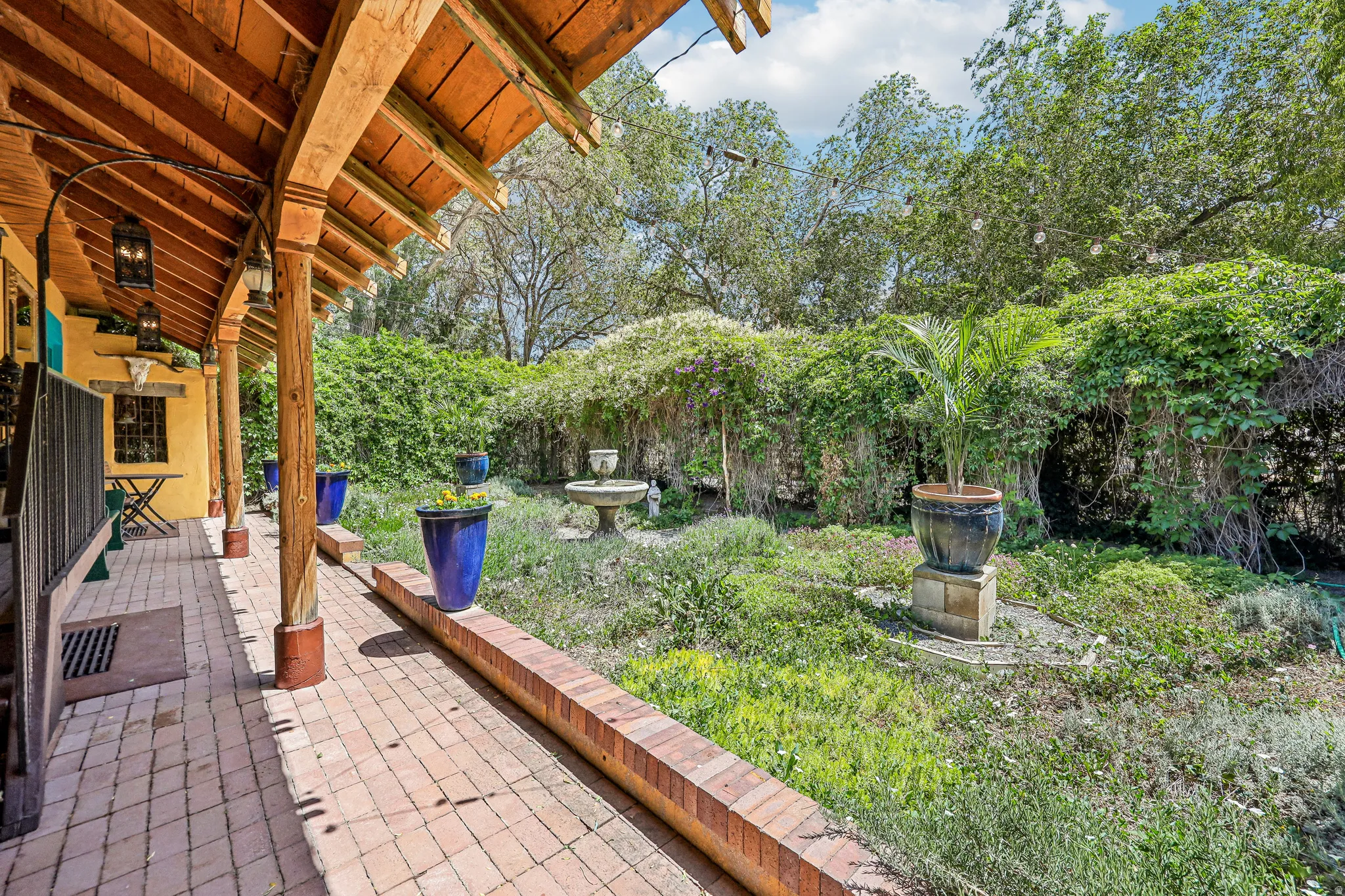 Covered patio with a brick paver floor, exposed wood beam ceiling, and decorative columns overlooking a lush garden with a central fountain