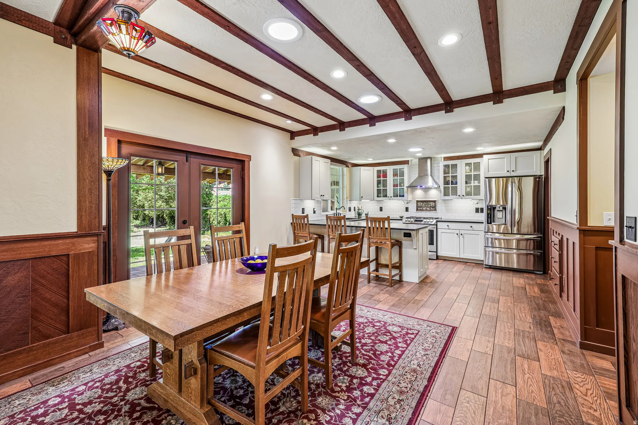 Open-concept kitchen and dining area featuring wood-finish flooring, white cabinetry, stainless steel appliances, and a beamed ceiling with recessed lighting
