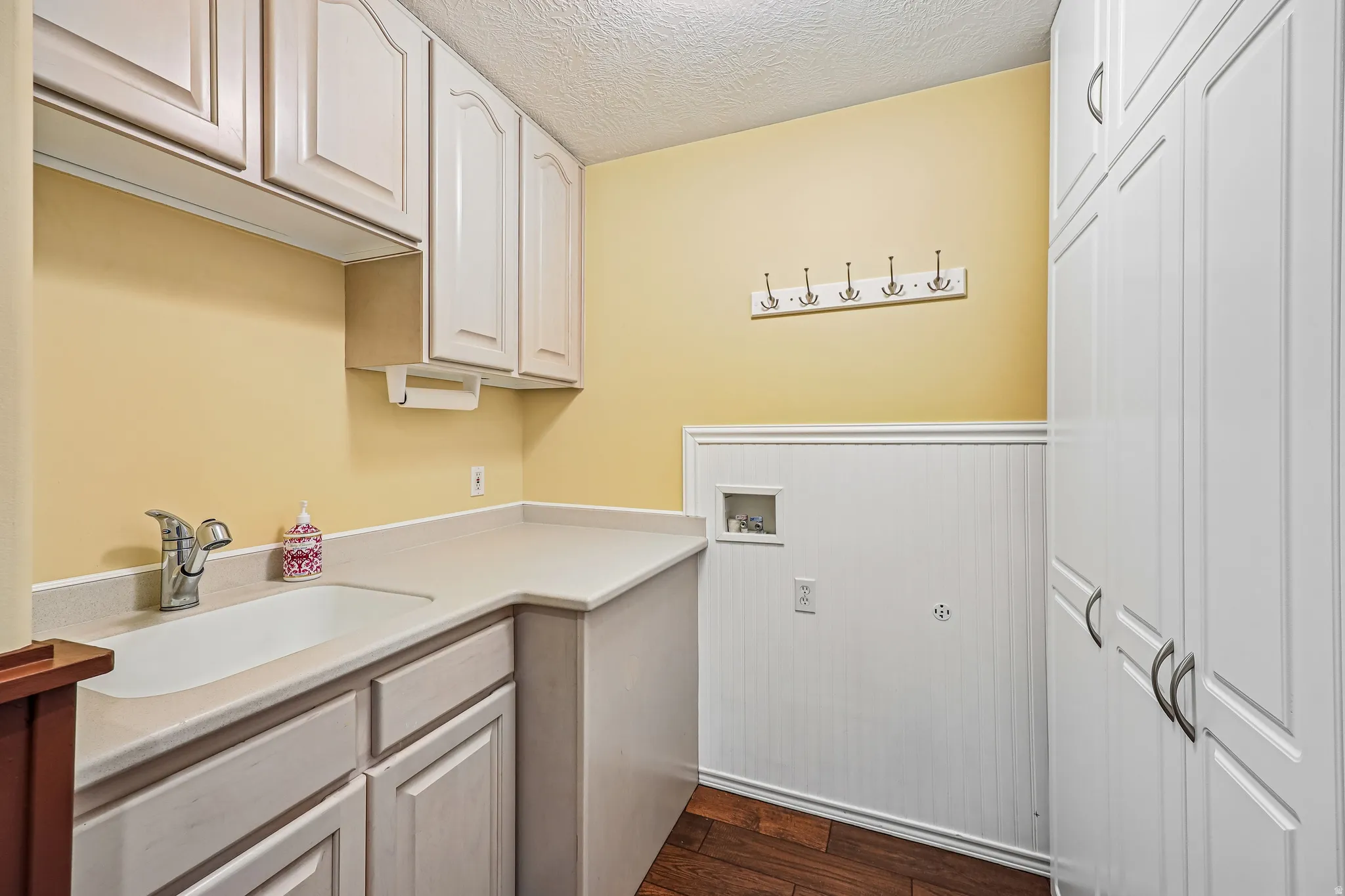 Utility room featuring extensive white cabinetry, a built-in sink with a chrome faucet, light-colored countertops, and rich wood-finish flooring
