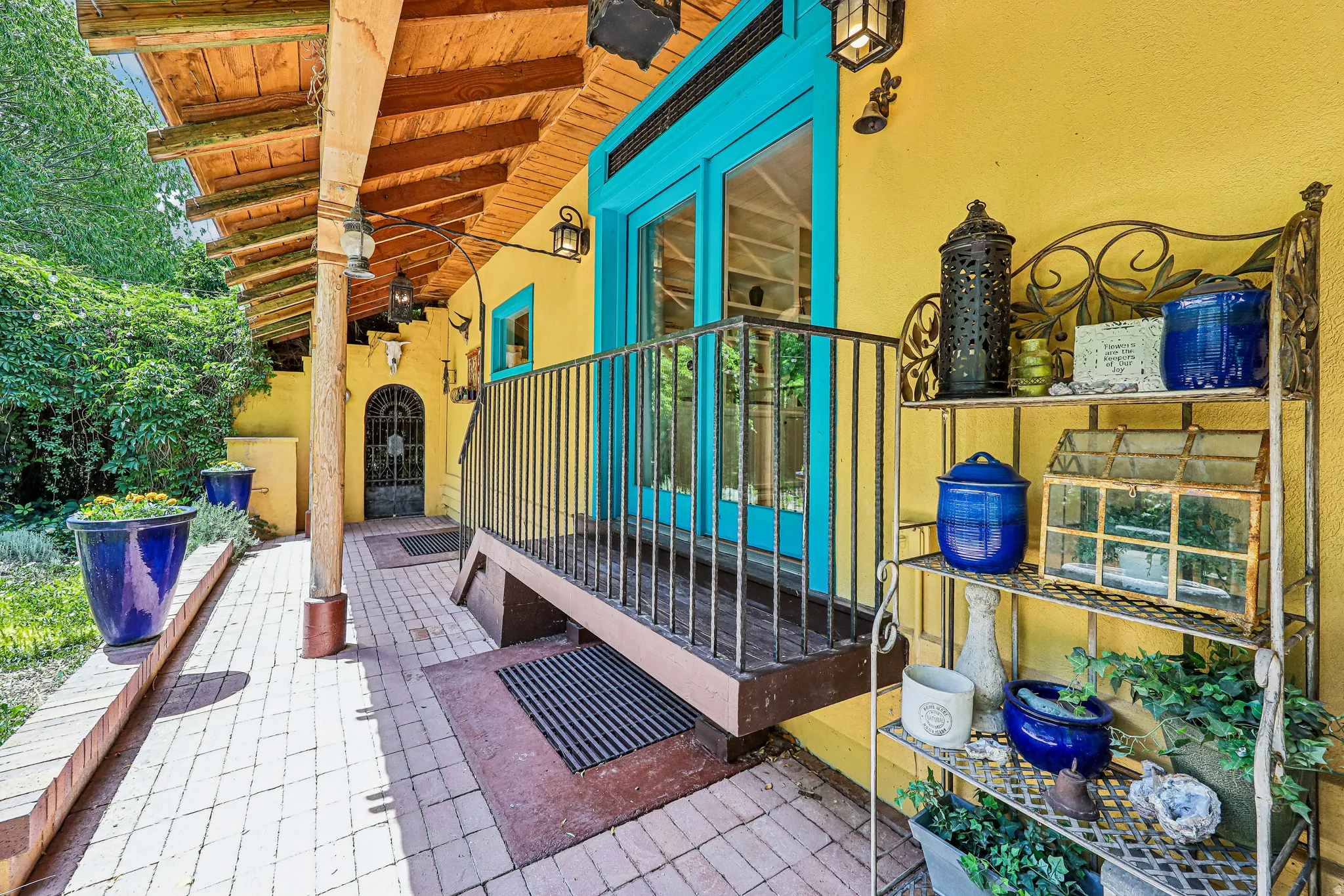 Vibrant yellow exterior featuring a covered porch with exposed wood beams, brick paver flooring, and bright turquoise double doors with wrought iron detailing
