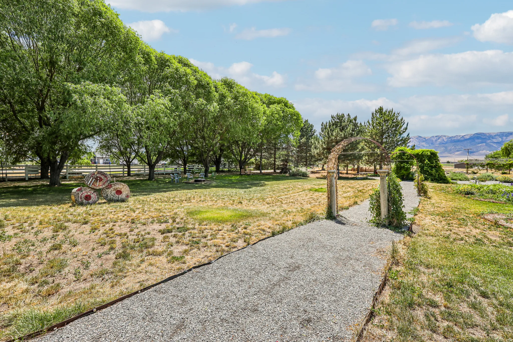 Expansive grounds featuring a gravel pathway, mature trees, white fencing, and distant mountain ranges