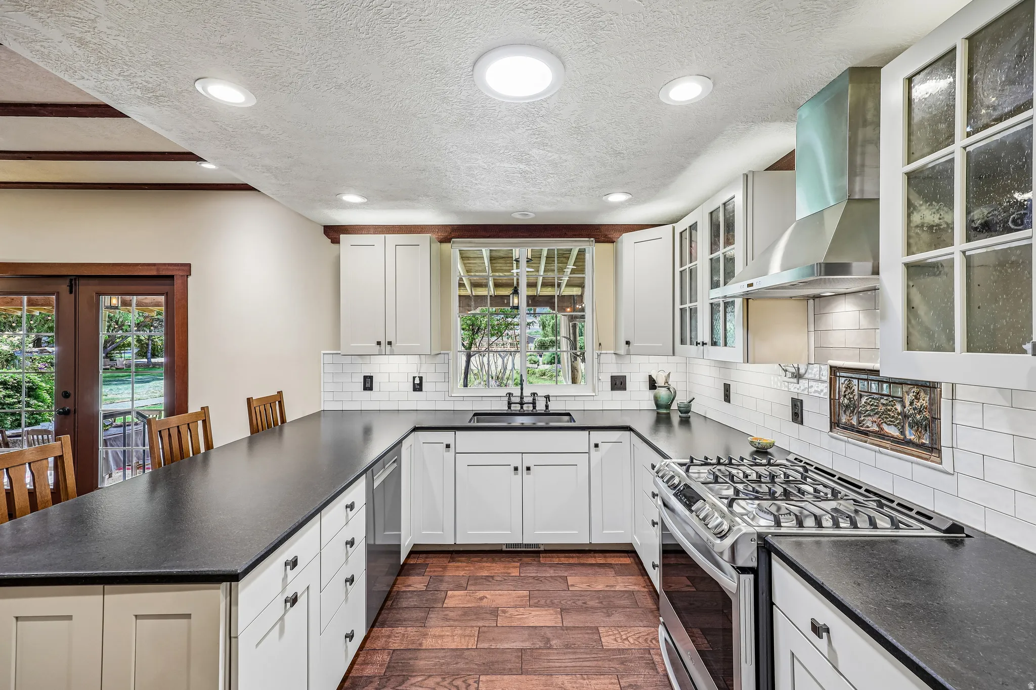U-shaped kitchen featuring white cabinetry, dark countertops, wood-finish flooring, a stainless steel gas range with a matching range hood, and a subway tile backsplash