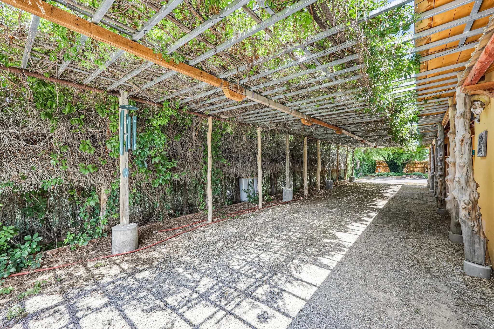 Covered walkway featuring a metal pergola structure with mature vine growth, gravel surfacing, and natural wood support posts