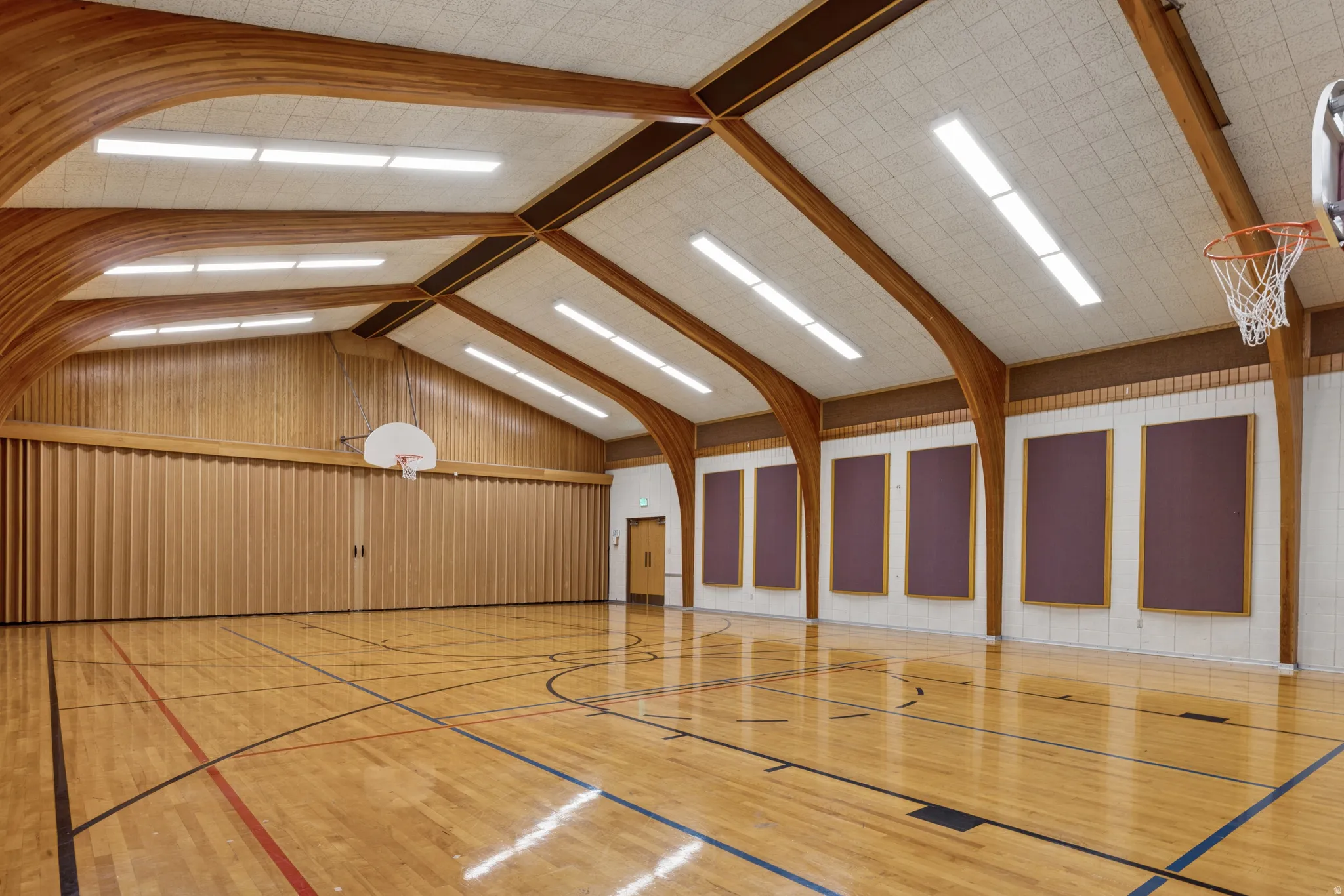Gymnasium space featuring wood-finish flooring with court markings, exposed wood beam ceiling, acoustic paneling, and multiple basketball hoops.