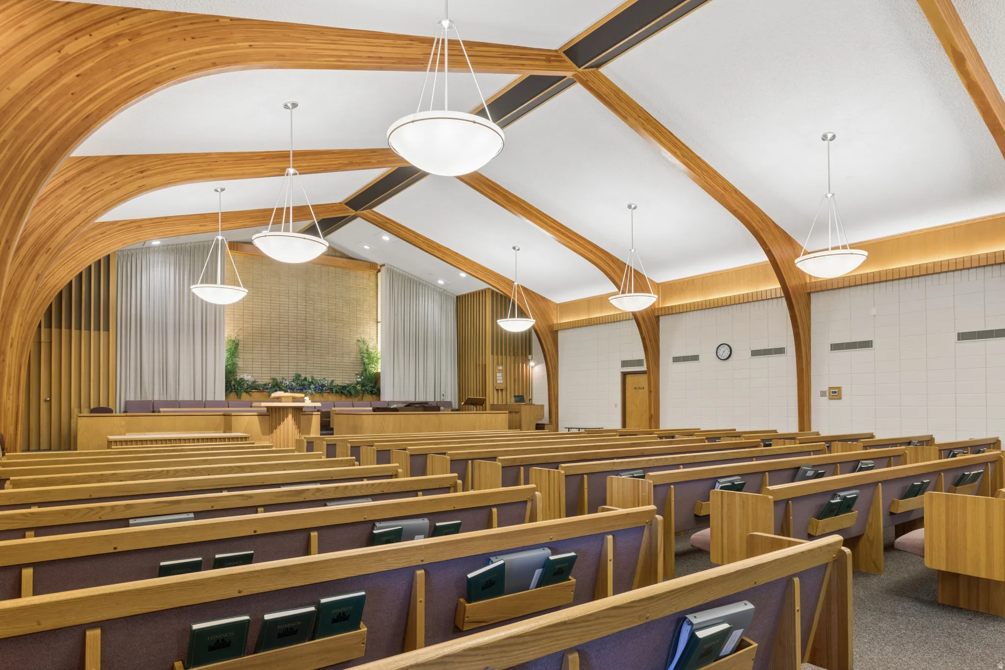 Spacious interior featuring a vaulted ceiling with exposed wood beams, multiple rows of wood seating with upholstered cushions, and contemporary pendant lighting fixtures.