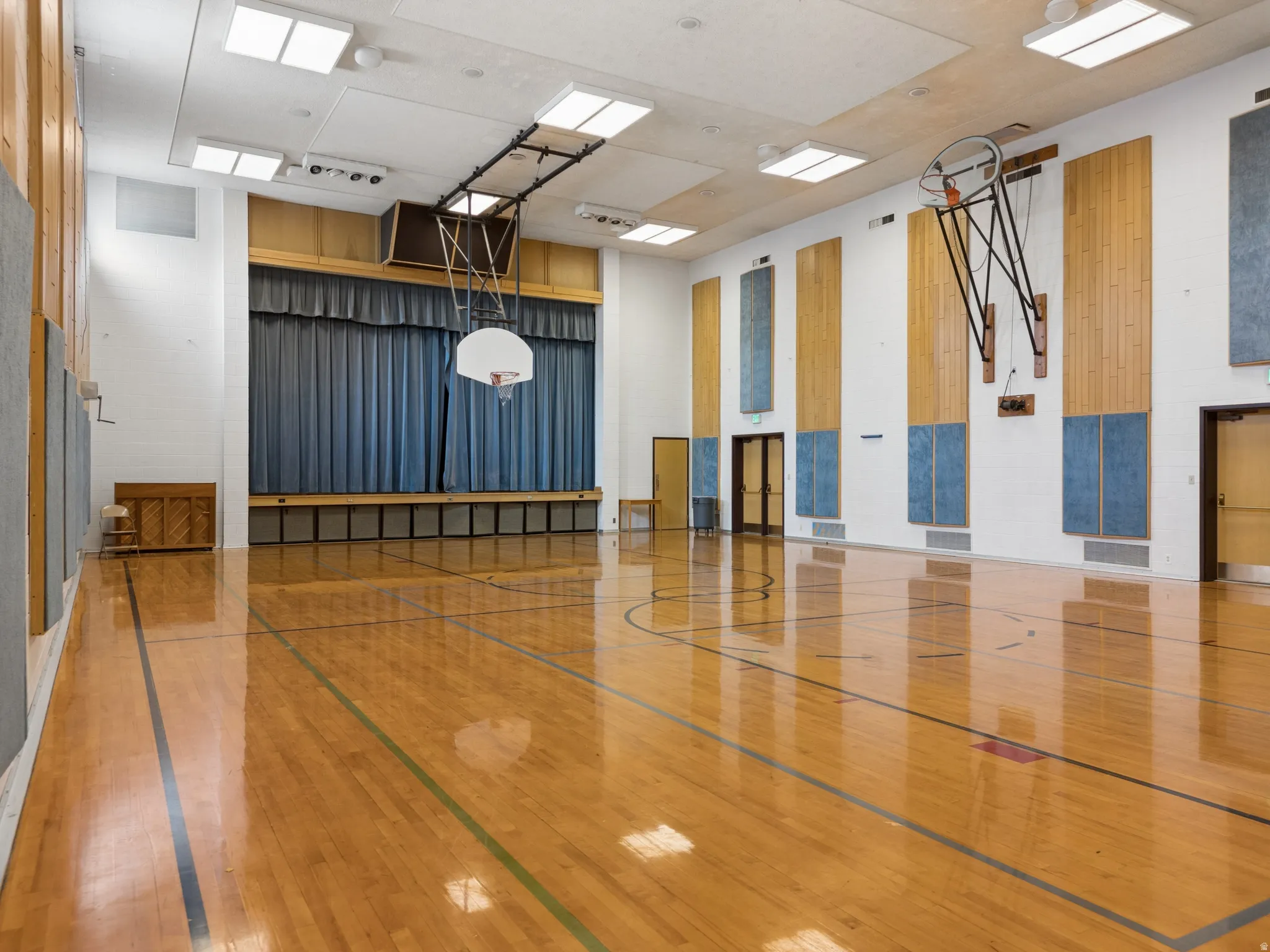 Spacious gymnasium featuring polished wood flooring with court lines, a built-in stage with curtain, two basketball hoops, wood-paneled wall accents, and recessed ceiling lighting.