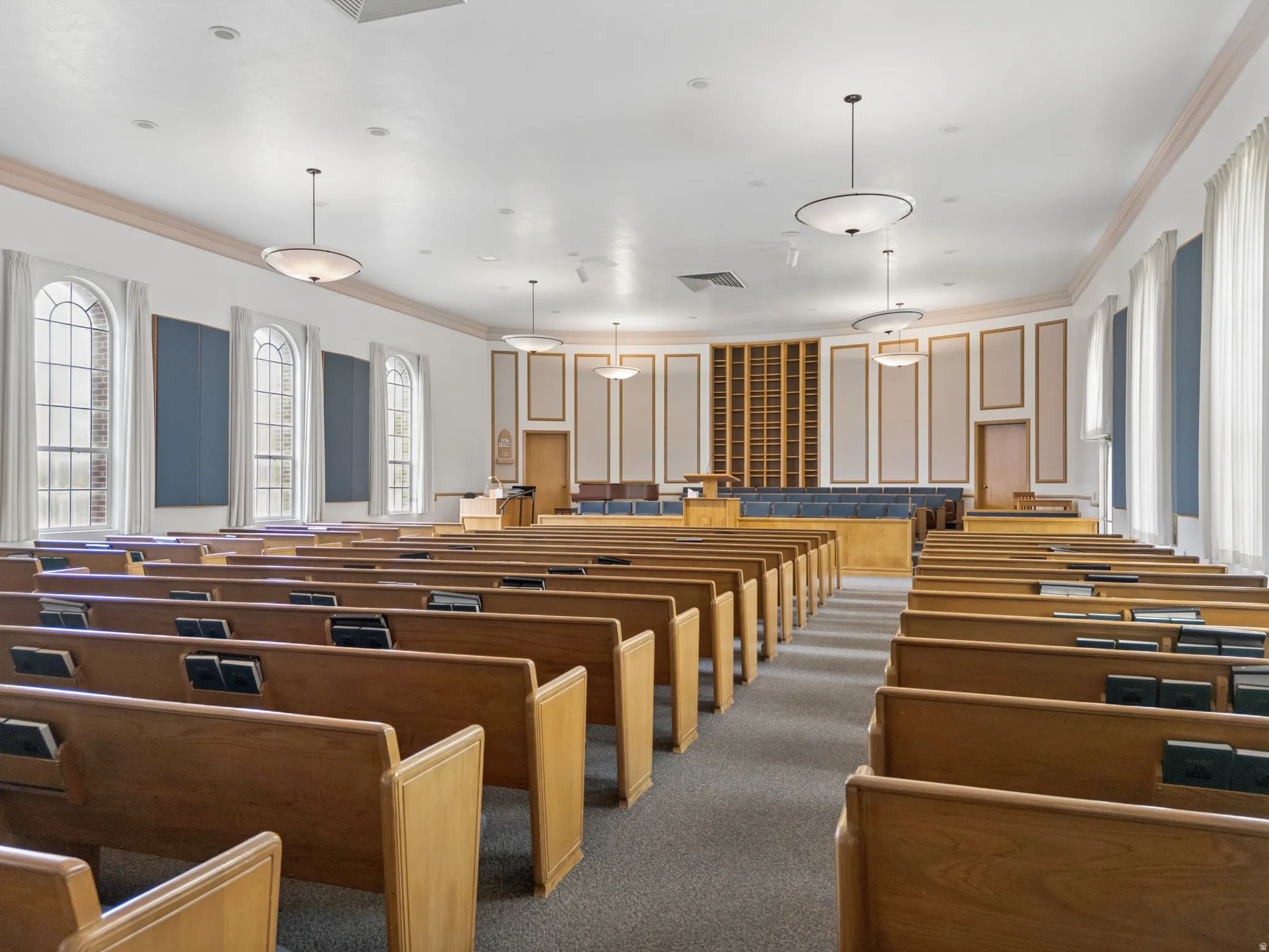 Spacious assembly hall featuring multiple rows of wooden pews, arched windows with white drapes, and recessed and pendant lighting.