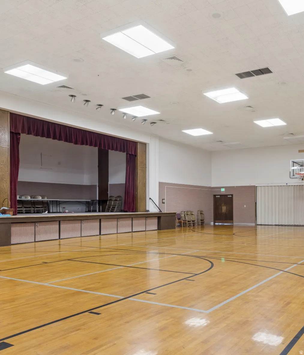 Hardwood-finish gymnasium flooring featuring court line markings, a raised stage with a curtain, recessed ceiling lighting, and a basketball hoop.