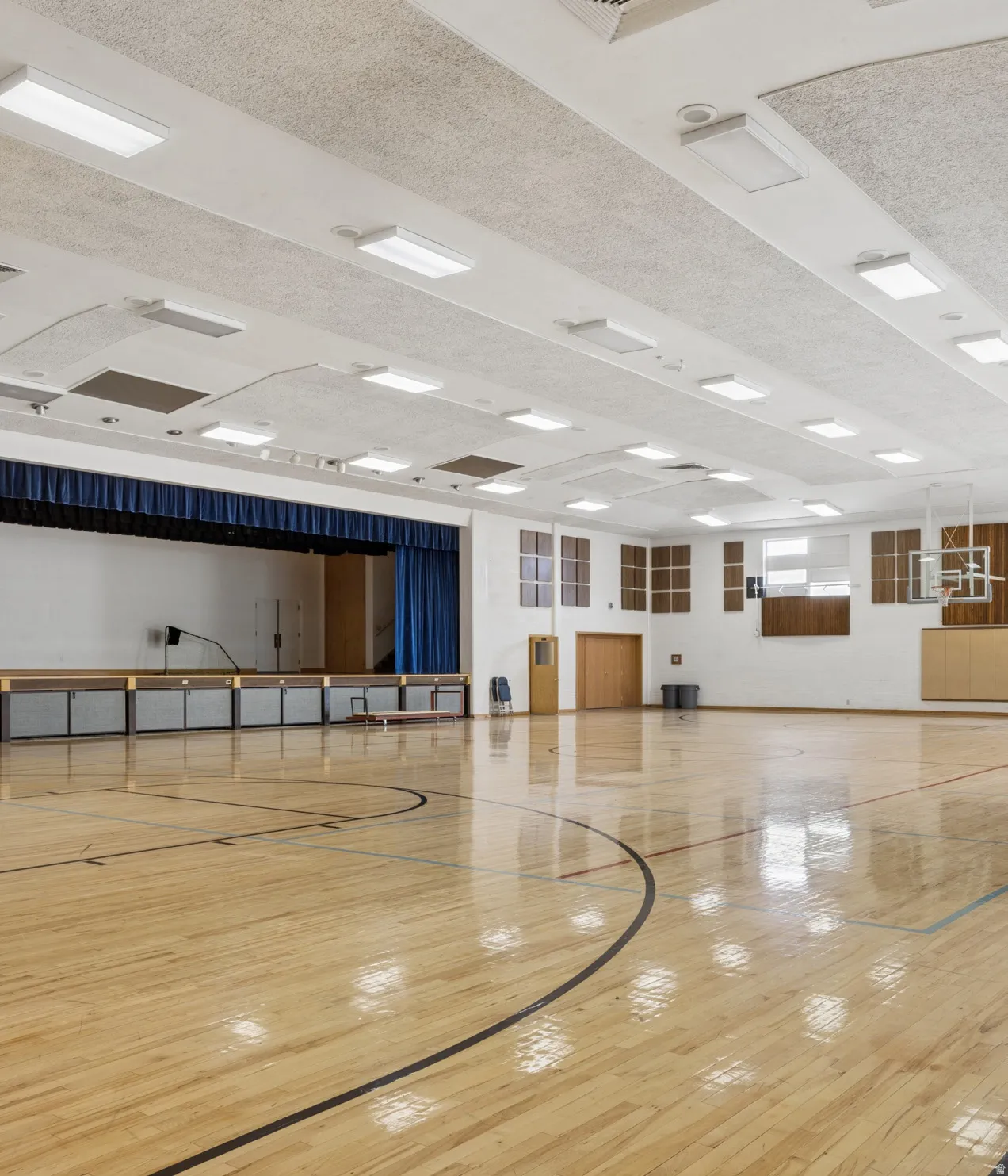 Spacious gymnasium featuring polished wood-finish flooring with court markings, a performance stage with blue curtains, overhead fluorescent lighting, acoustic ceiling panels, and multiple wall-mounted windows.