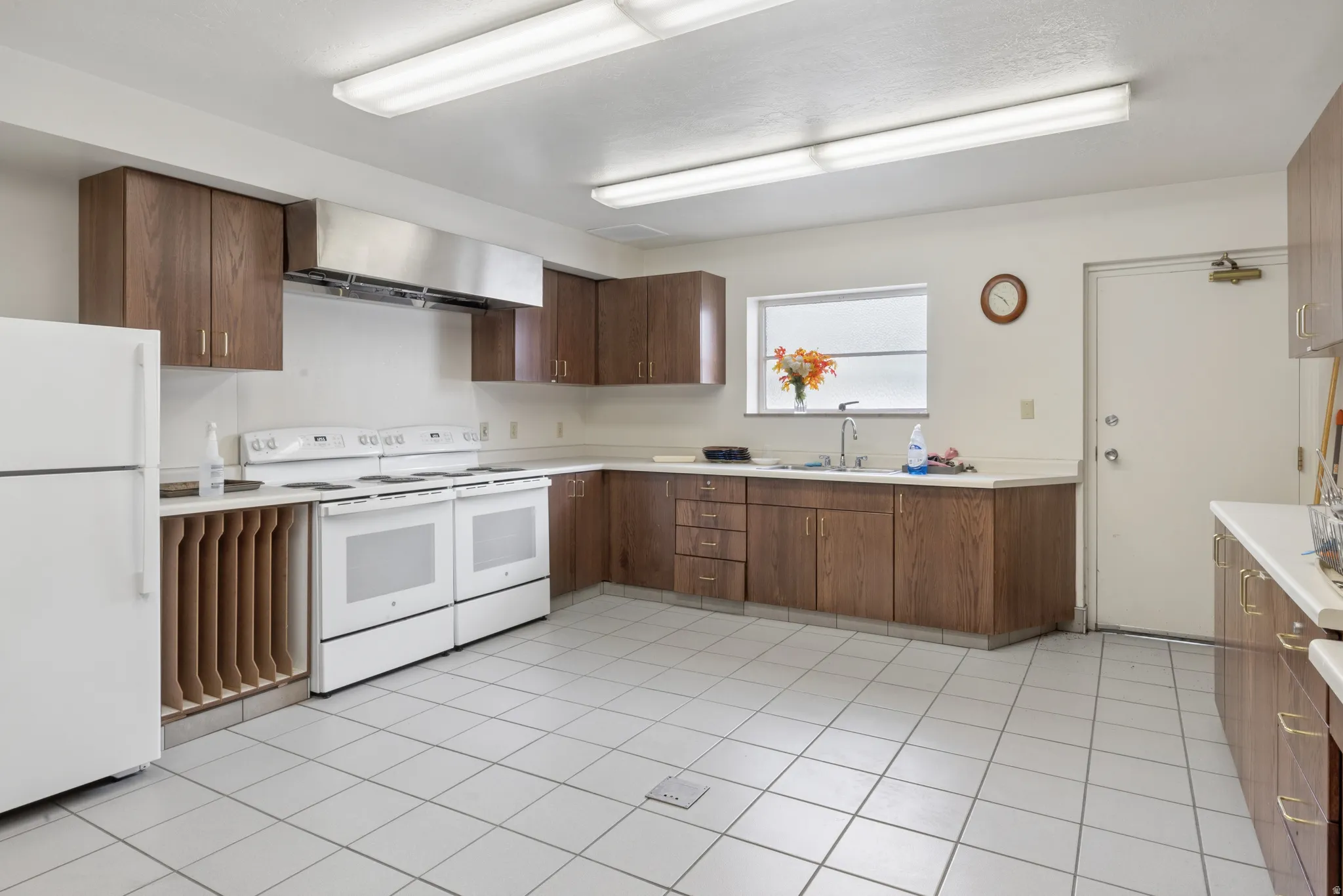 Expansive kitchen featuring white square floor tiles, wood-finish cabinetry, a large window, a stainless steel range hood, and multiple white appliances.