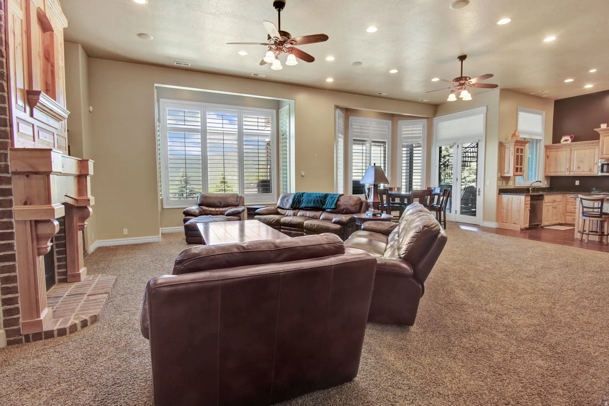 Spacious open-concept living area featuring a brick fireplace with a wood mantel, large windows with white louvered shutters, recessed lighting, and ceiling fans