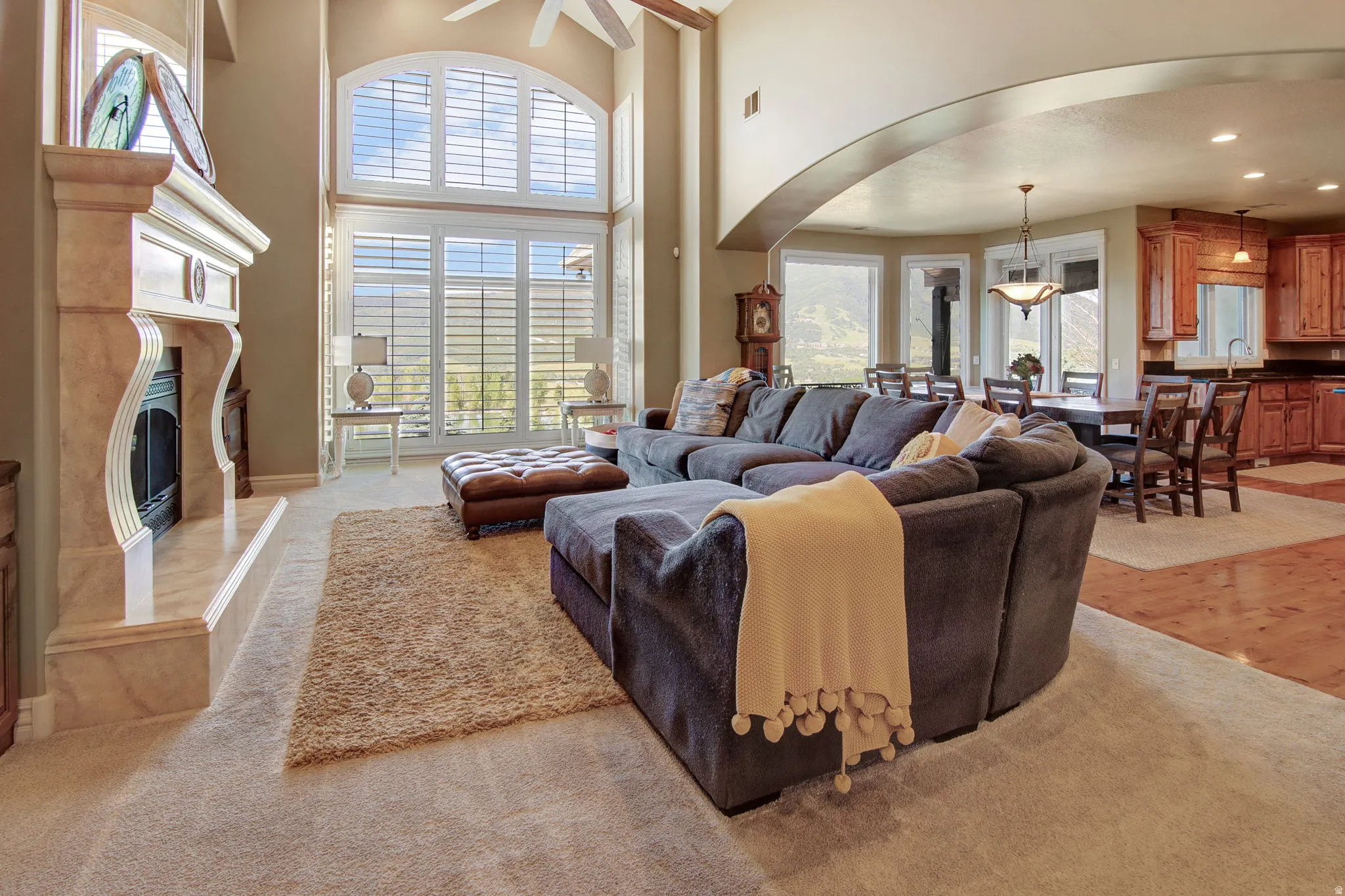 Living area featuring a floor-to-ceiling window array with plantation shutters, a fireplace with a stone mantel, vaulted ceilings, and an arched entry to a dining space with wood-finish flooring