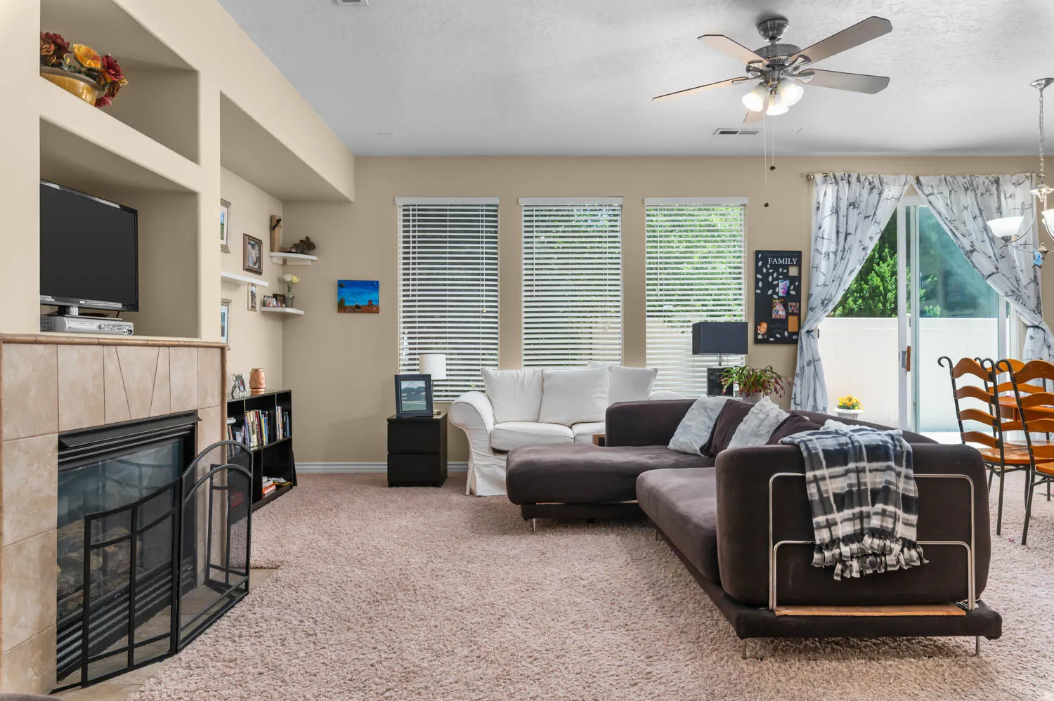 Living area featuring a tile-surround fireplace with built-in shelving, carpeted flooring, three large windows with blinds, a ceiling fan with integrated lighting, and a glass sliding door
