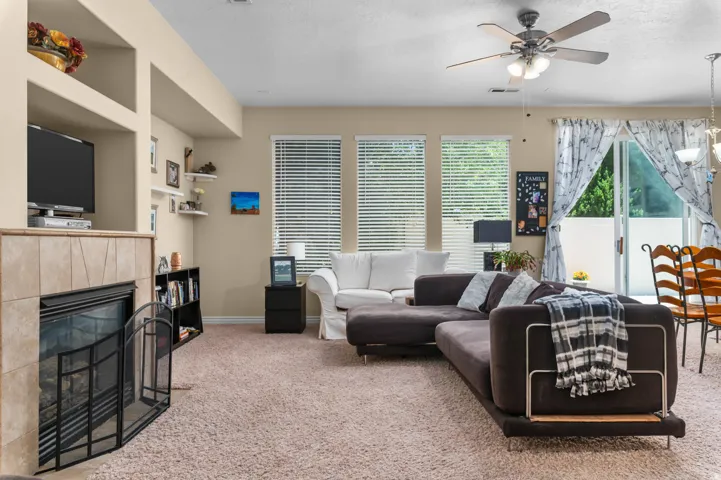 Living area featuring a tile-surround fireplace with built-in shelving, carpeted flooring, three large windows with blinds, a ceiling fan with integrated lighting, and a glass sliding door