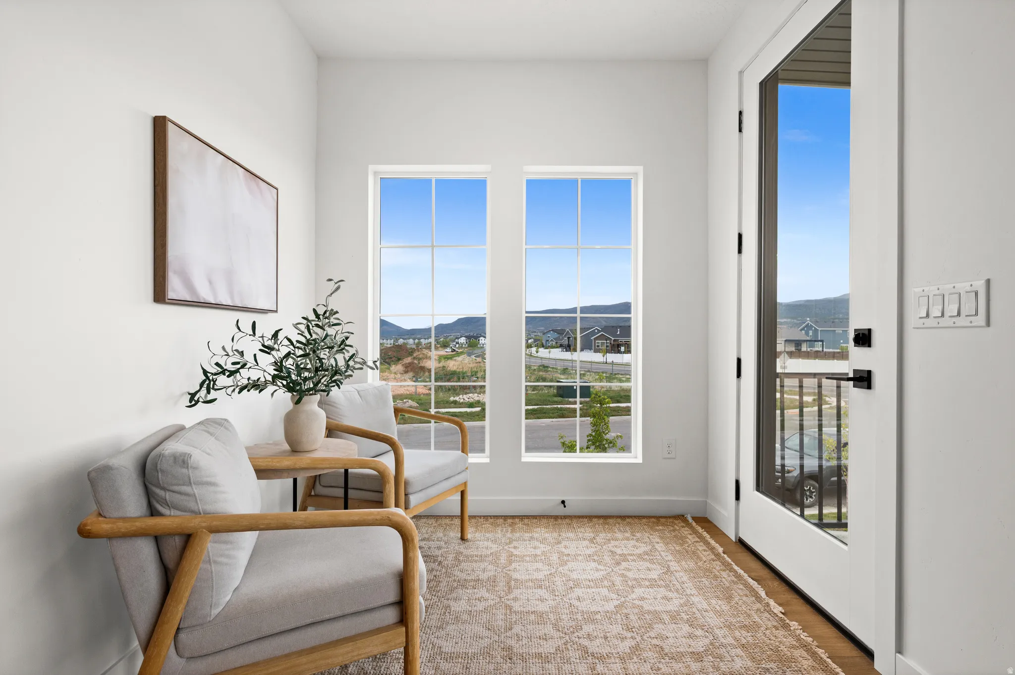 Bright interior space featuring a full-light exterior door with matte black hardware, two large multi-pane windows, wood-finish flooring, and crisp white walls