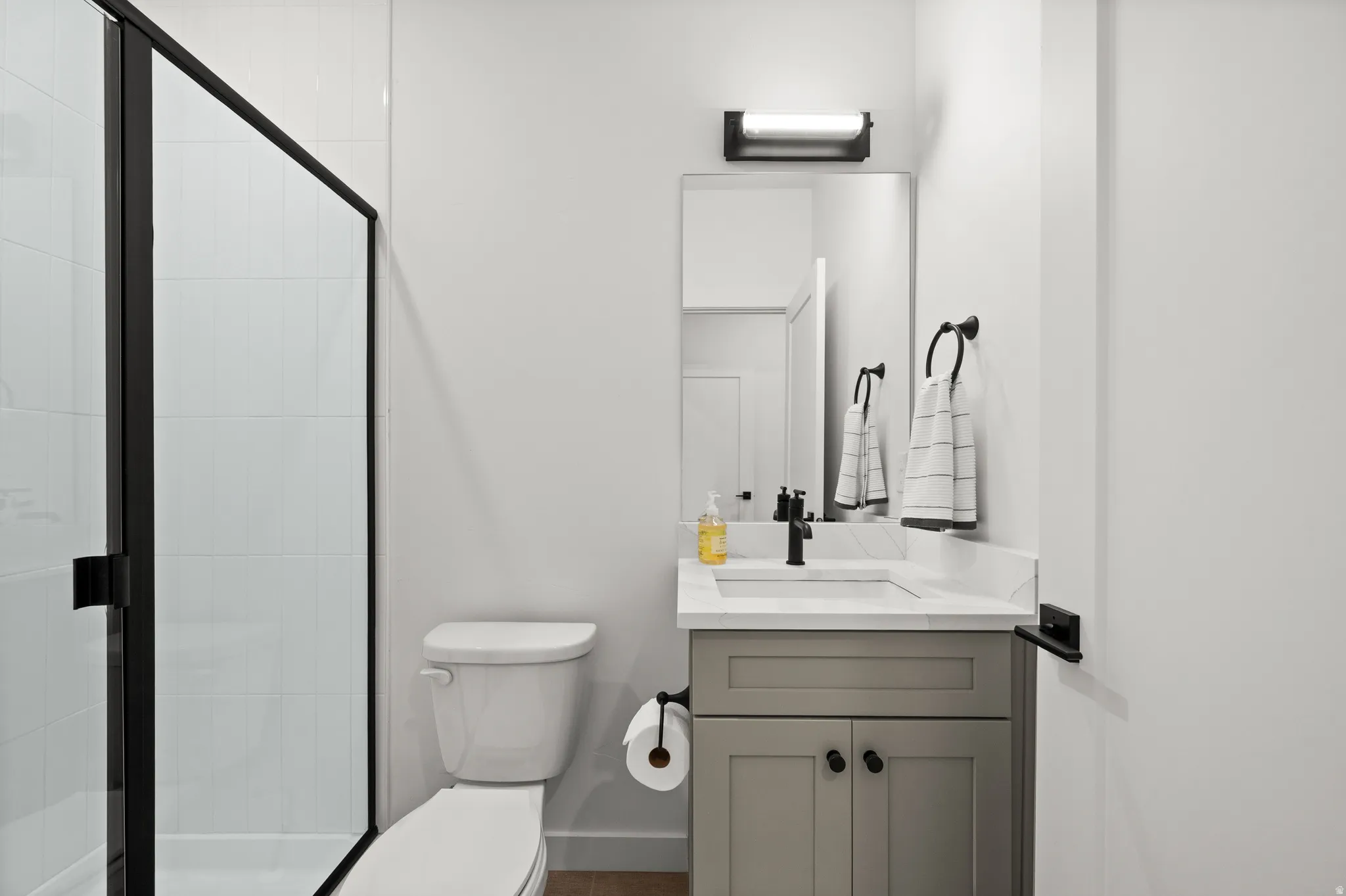 Bathroom featuring a glass-enclosed shower with black trim, a vanity with a grey cabinet and white countertop, an undermount sink with a matte black faucet, a rectangular mirror, and a wall-mounted light fixture