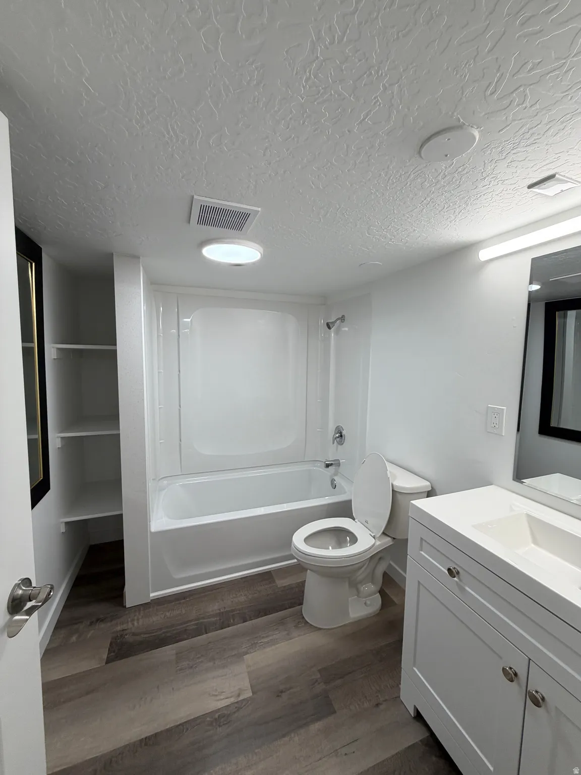 Bathroom featuring wood-finish flooring, a white vanity with a single basin sink, a white bathtub with a shower surround, and integrated shelving