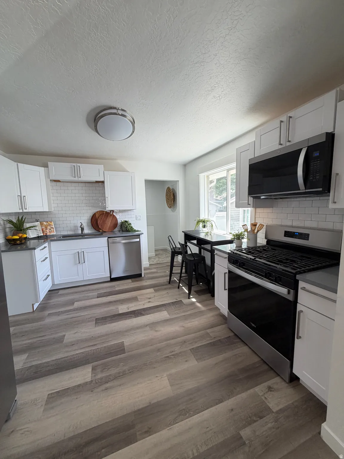 Kitchen featuring wood-finish plank flooring, white shaker cabinetry, stainless steel appliances, dark countertops, and a white subway tile backsplash