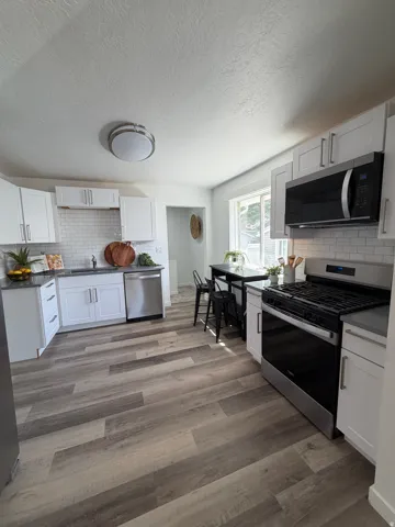 Kitchen featuring wood-finish plank flooring, white shaker cabinetry, stainless steel appliances, dark countertops, and a white subway tile backsplash
