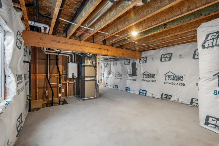 Unfinished basement space featuring exposed wood joists and subfloor, concrete flooring, window opening, and visible utility connections including a water heater and furnace