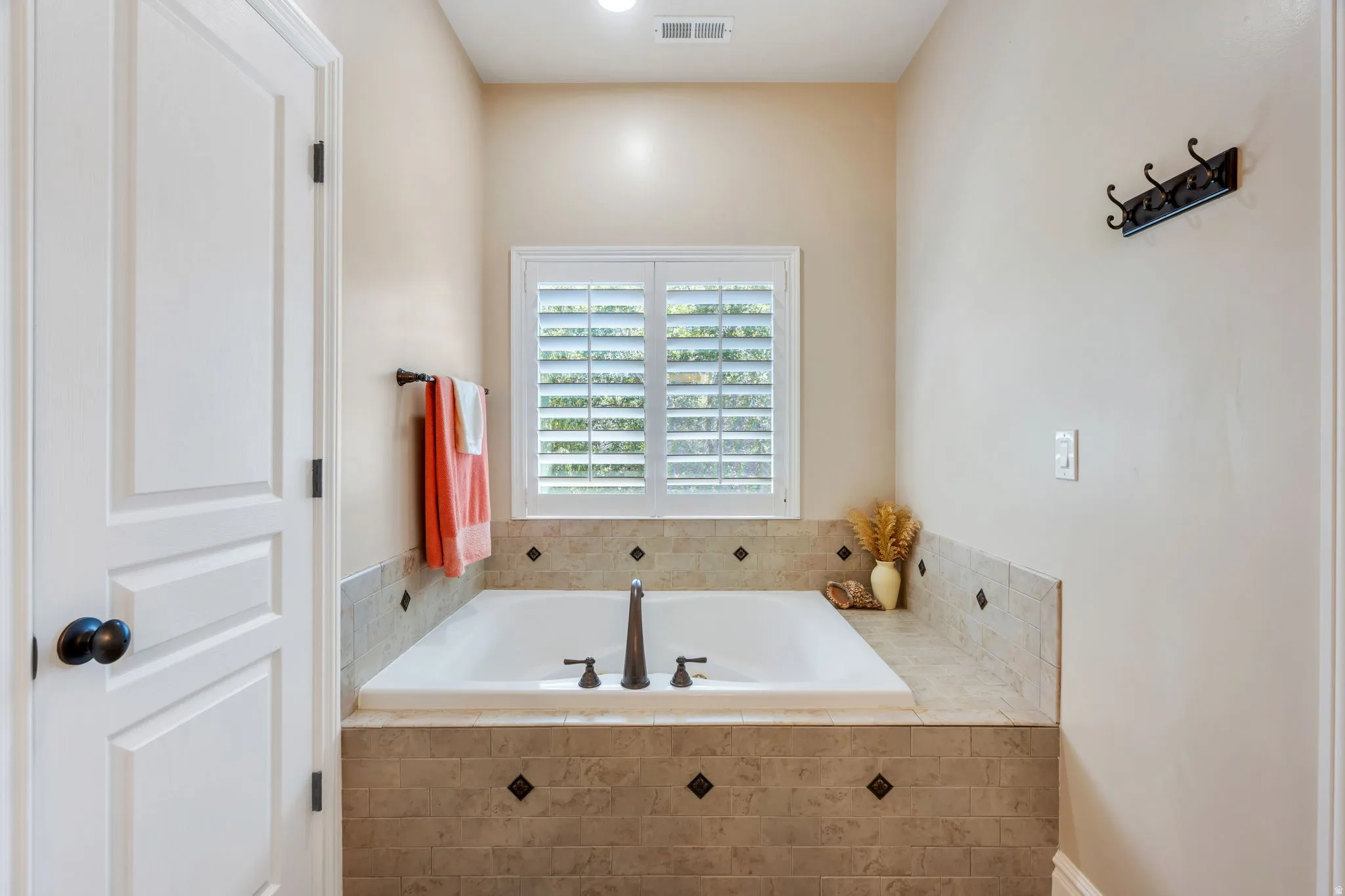 Built-in soaking tub with a tiled surround featuring dark accent squares, a window with plantation shutters, and oil-rubbed bronze fixtures
