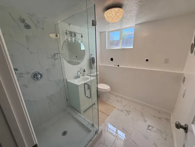 Modern bathroom featuring a glass-enclosed shower with white marble-look tile, a white vanity with a rectangular sink, a circular mirror, and white marble-look flooring