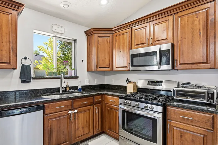 Kitchen with wood cabinetry, dark countertops, stainless steel appliances, and a double basin sink beneath a window