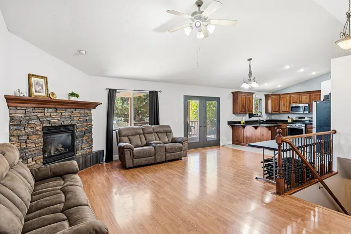 Open-concept interior featuring wood-finish flooring, a stone-faced fireplace with a wood mantel, vaulted ceilings with a ceiling fan, and French doors leading to exterior space
