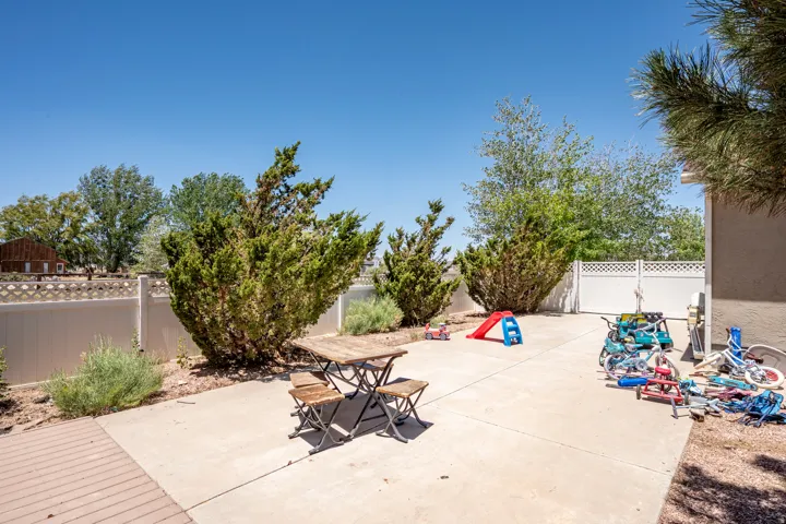 Spacious concrete patio area featuring a white perimeter fence, mature shrubs, and an adjacent wood-finish deck section