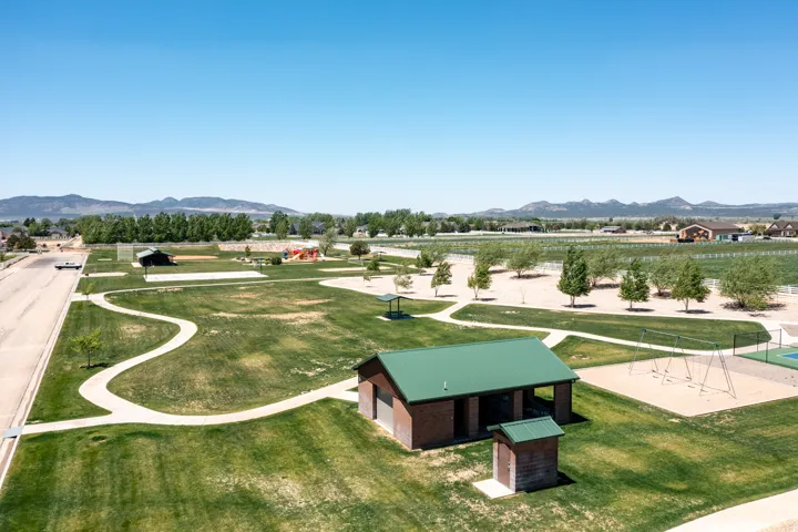 Expansive green space featuring a winding paved pathway, a brick pavilion with a green metal roof, a sand-based play area with swing sets, and a hard-surface court