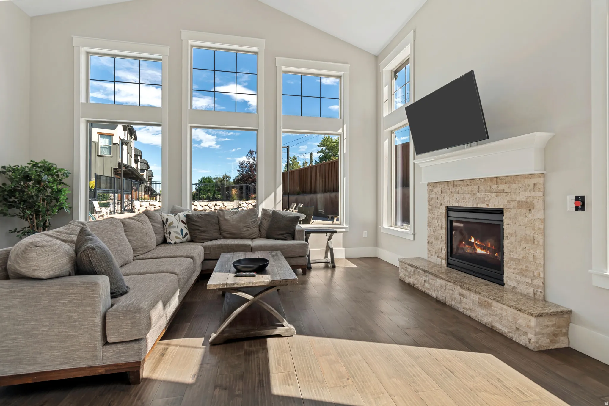 Vaulted ceiling, wood-finish flooring, stone-clad fireplace, white mantel, and multiple large windows