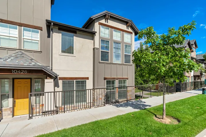 Contemporary facade with stucco and wood-look siding, multi-pane windows with blinds, a prominent yellow front door, a sloped shingle entryway, and a low iron fence