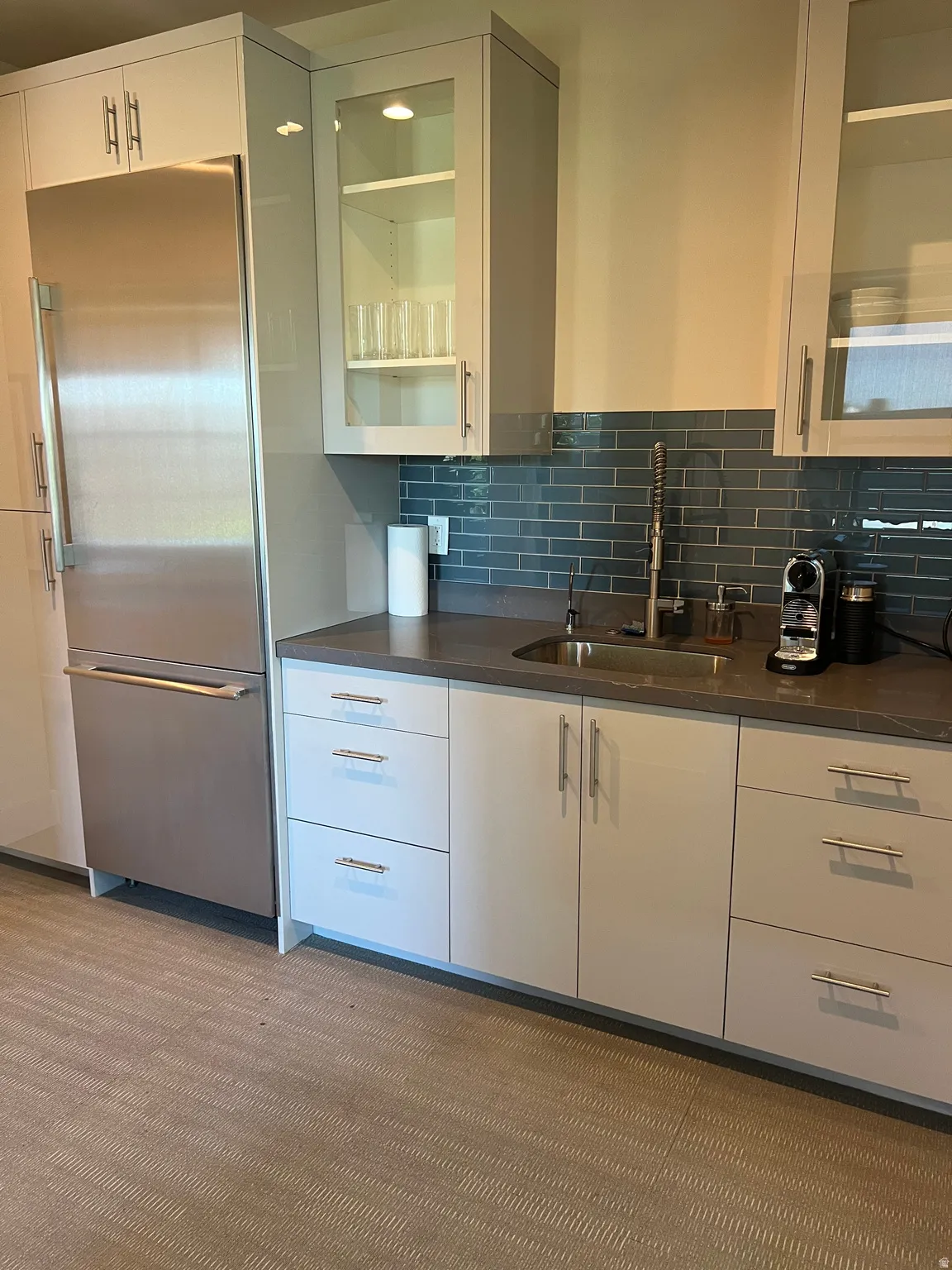 Kitchenette featuring a stainless steel refrigerator, white cabinetry with polished hardware, dark stone-finish countertops, an undermount sink, and a blue subway tile backsplash