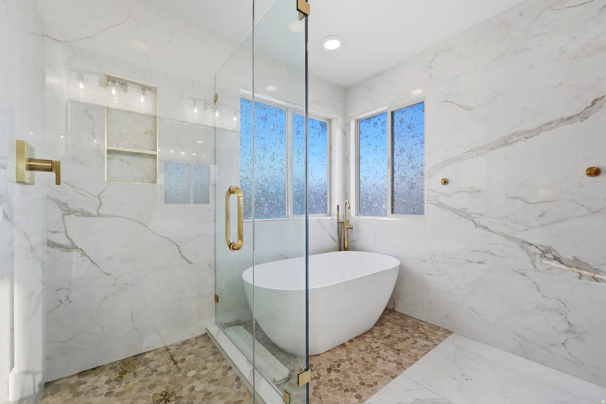 Spa-like bathroom featuring a freestanding soaking tub, glass-enclosed shower, gold-toned fixtures, marble-look wall tiling, and pebble-style flooring