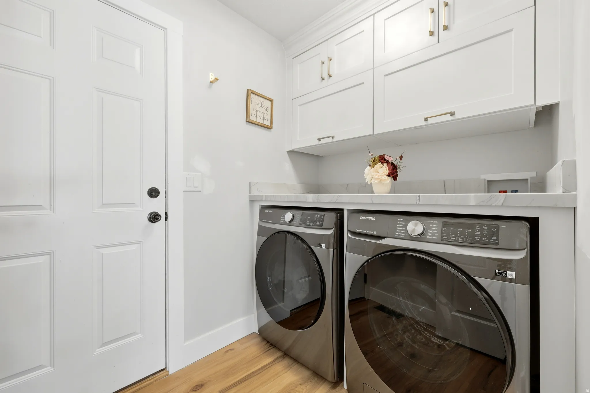 Dedicated laundry area featuring wood-finish flooring, white paneled cabinetry with brass hardware, and a light-toned countertop
