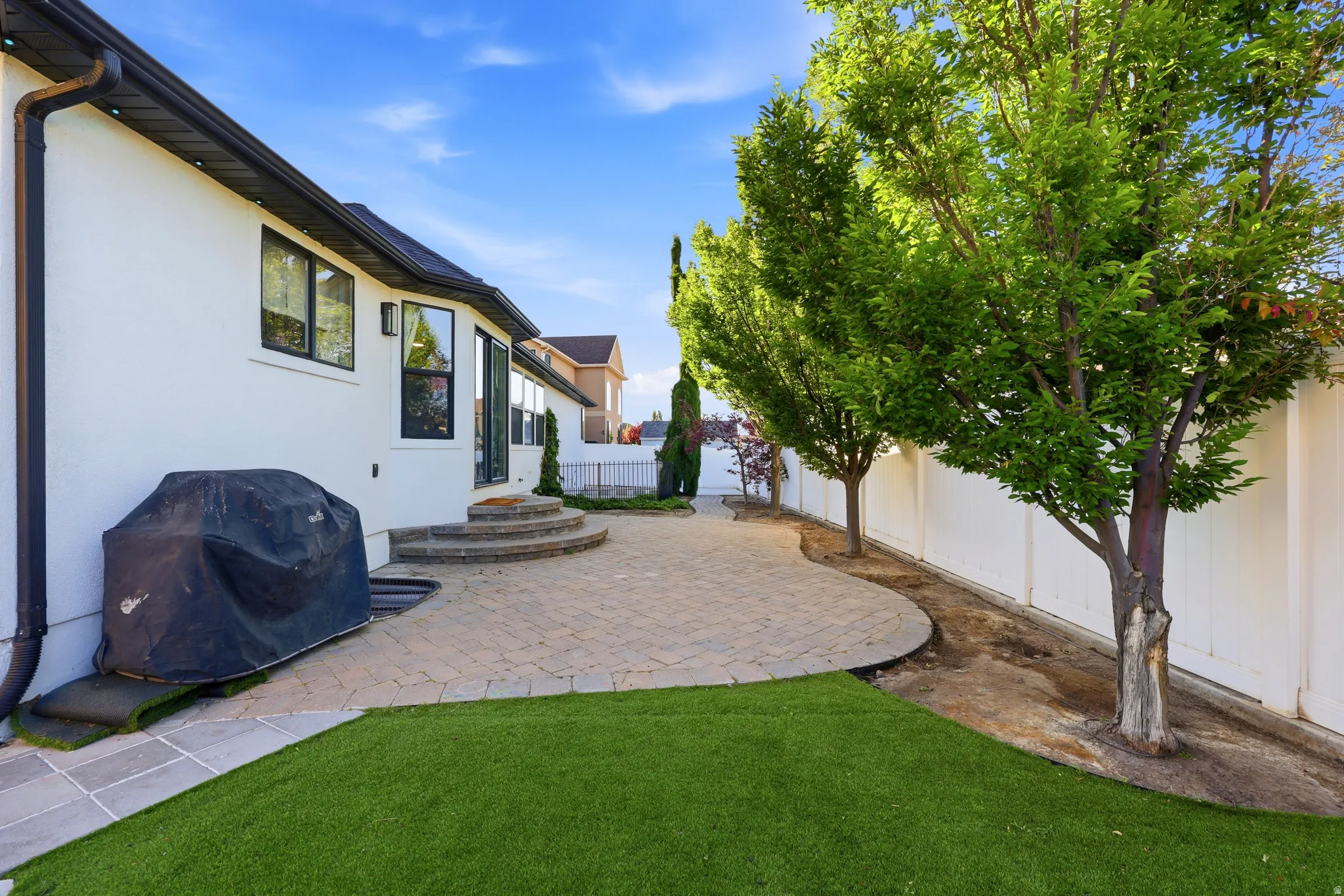 Outdoor living area featuring a paver patio with curved steps, a white stucco exterior, black-framed windows, and mature trees lining a privacy fence