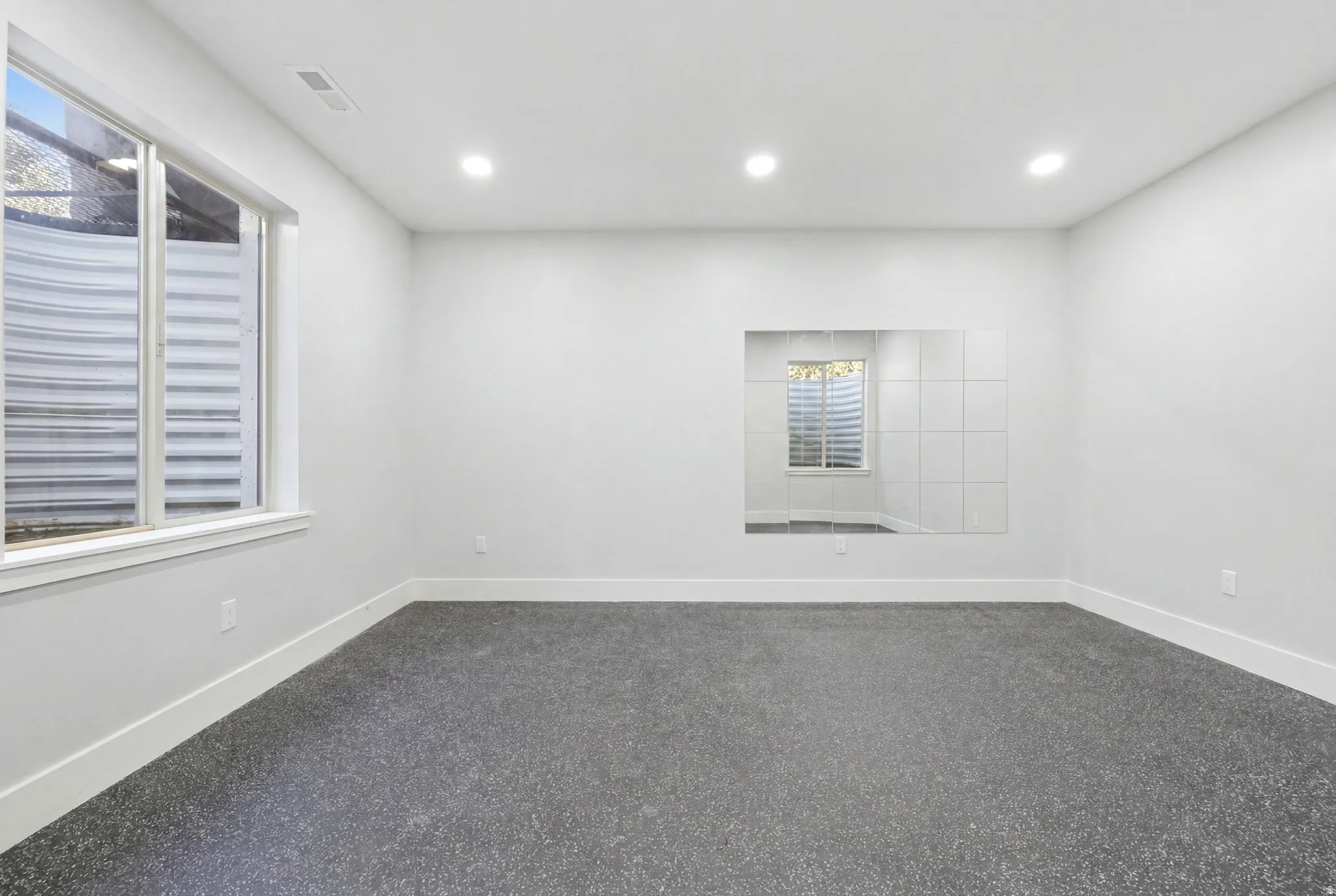 Spacious room featuring light gray walls, recessed ceiling lighting, a large window with a window well, dark speckled flooring, and a mirrored wall section