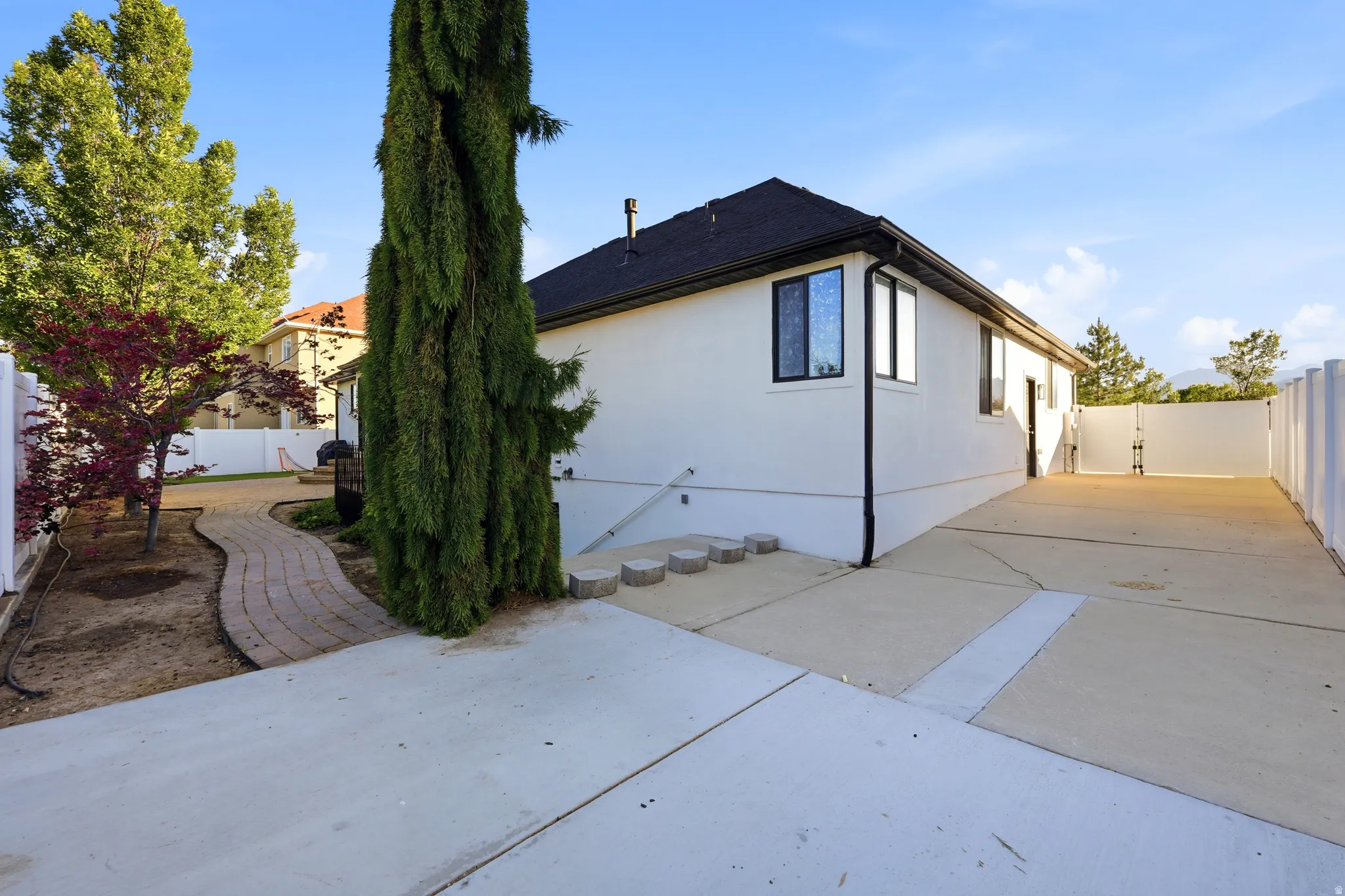 Expansive concrete paving alongside the property, featuring a white stucco exterior with black-framed windows, a dark shingled roof, and a white privacy fence