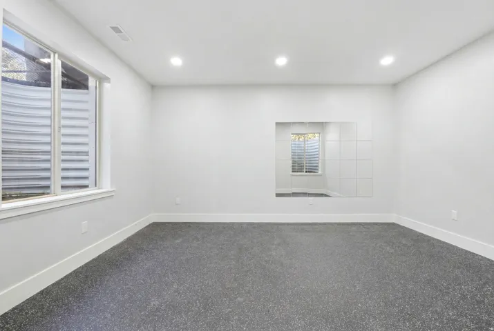Spacious room featuring light gray walls, recessed ceiling lighting, a large window with a window well, dark speckled flooring, and a mirrored wall section