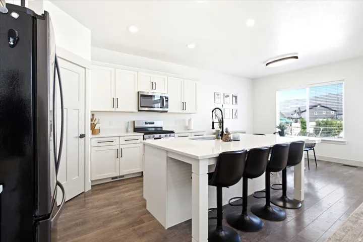 Open-concept kitchen featuring white shaker cabinetry, stainless steel appliances, a center island with sink and seating, wood-finish flooring, and ample natural light from a large window