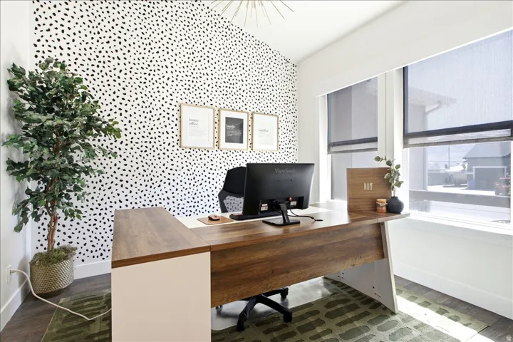 Office space featuring a patterned accent wall, large windows with roller shades, and a wood-finish L-shaped desk