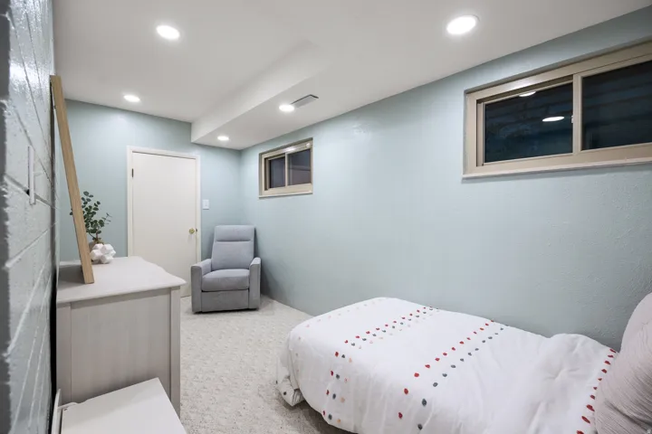 Recessed ceiling lighting illuminates a room featuring light blue walls, a white painted door with brass hardware, and a window with a tan frame