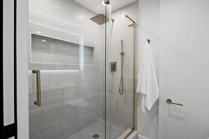 Walk-in shower featuring a frameless glass enclosure, a built-in shower niche with accent lighting, a rain showerhead, a hand-held shower fixture, and hexagonal floor tiling